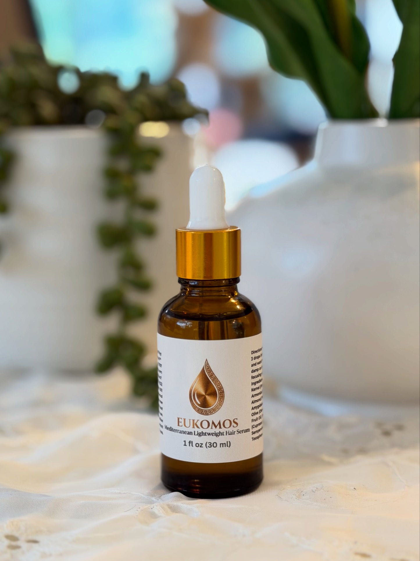 A 1 fl oz amber glass dropper bottle of Eukomos Light Night Sun essential oil, featuring a white label and a gold collar, set on a white lace cloth with blurred potted plants in the background.